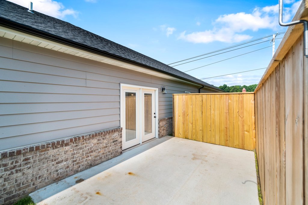 Side view of a house with gray siding, brick base, and double doors leading to a concrete patio and wooden fence.
