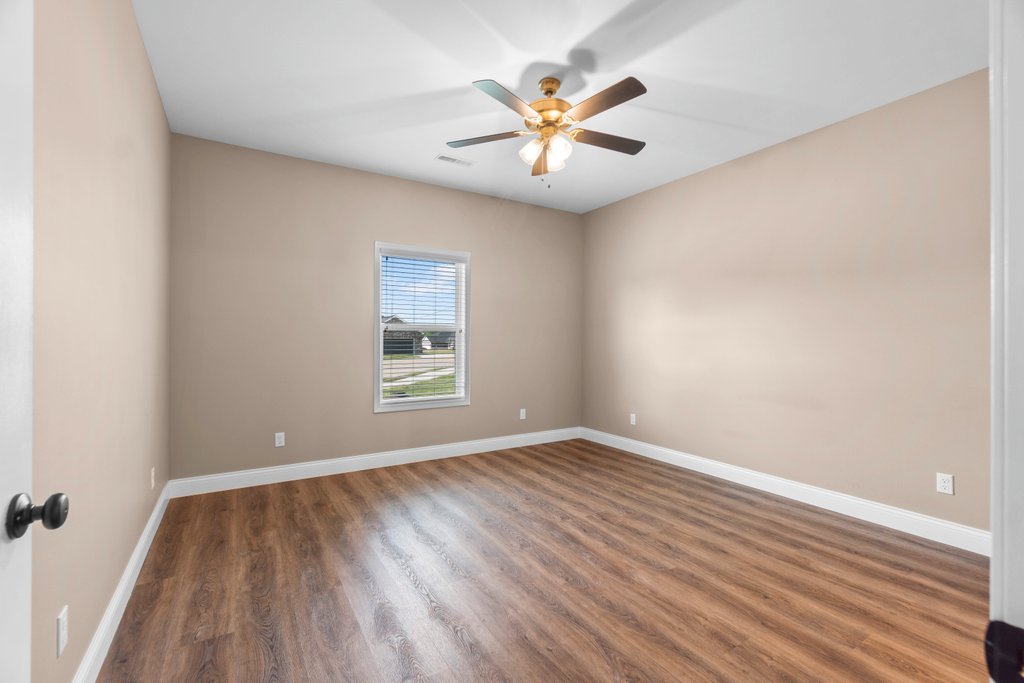 Empty beige bedroom with wood floors, ceiling fan, and window overlooking a suburban street.