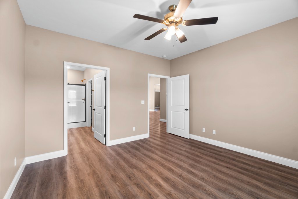 Modern bedroom with beige walls, wood floor, ceiling fan, and doors leading to a shower and hallway.