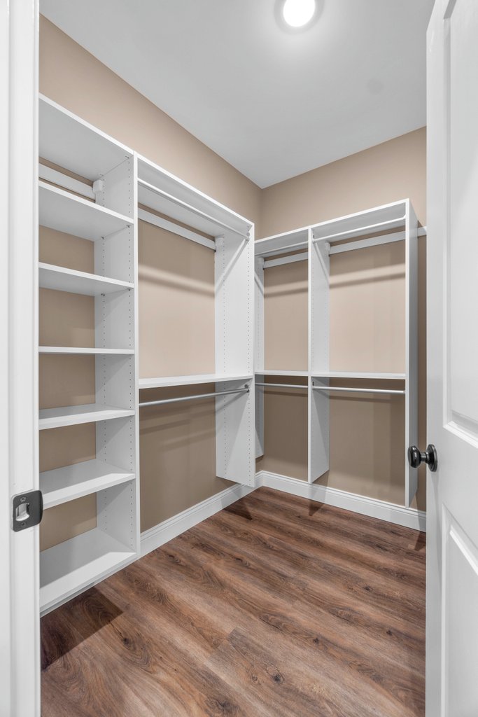 Empty walk-in closet with white shelves and rods, beige walls, and wood floor.