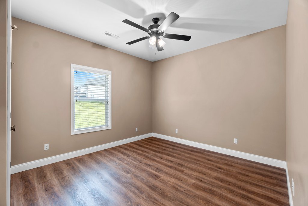 Empty bedroom with beige walls, wood floor, window, and ceiling fan.