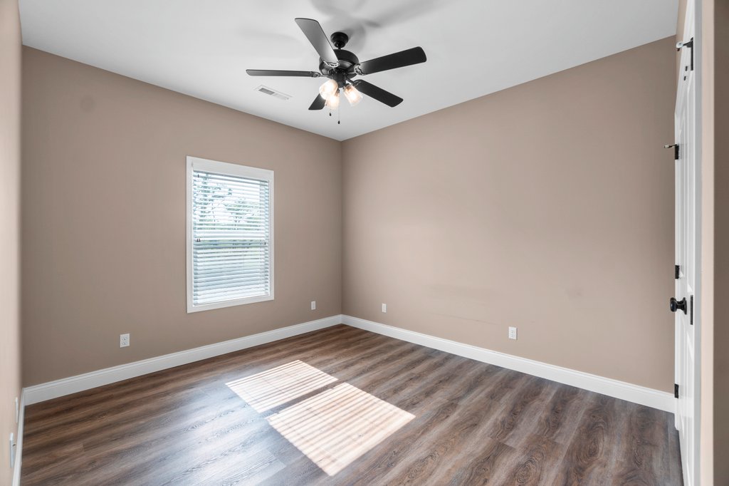 Empty bedroom with tan walls, wood floors, a ceiling fan, and a window with blinds.